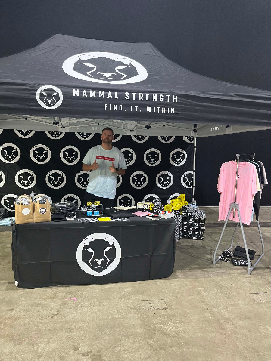 Person standing behind a table under a Mammal Strength tent with branded merchandise.